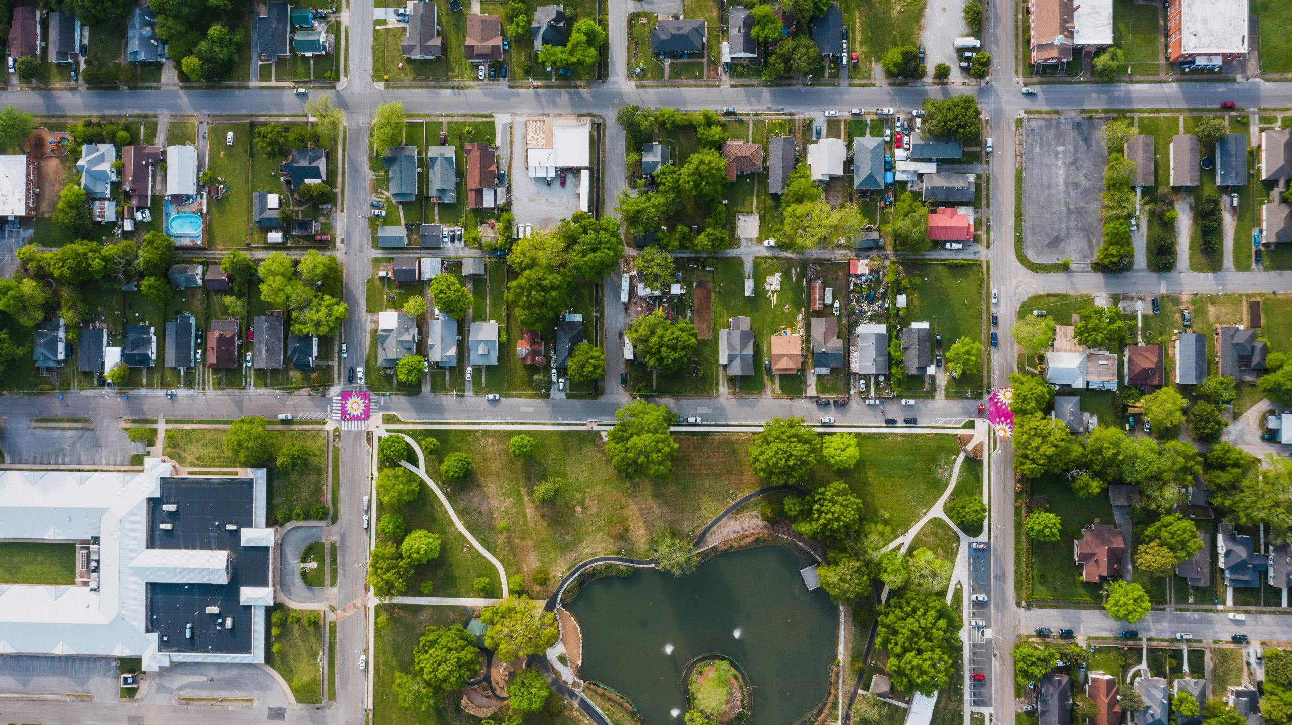 High-angle shot of a suburban neighborhood with streets, houses, and a park with a pond, showcasing vibrant greenery.